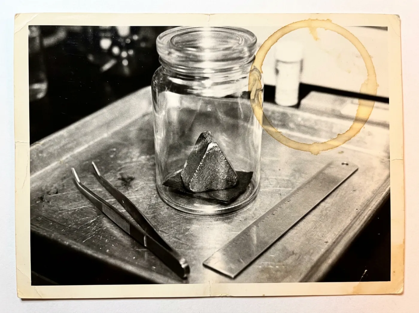Grainy black-and-white evidence photo of a small triangular metal sample in a glass jar on a lab tray, beside tweezers and a ruler, with a coffee ring stain on the print.